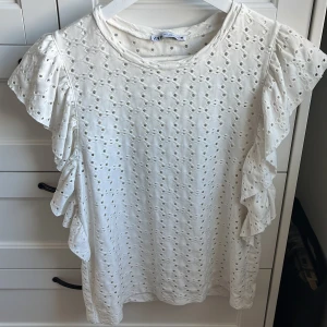 Vit blus med volangärmar från Zara - Säljer en söt vit blus från Zara med broderat hålmönster och volangdetaljer längs ärmarna. Blusen har rund halsringning och är tillverkad i bomull, perfekt för ljusa dagar.