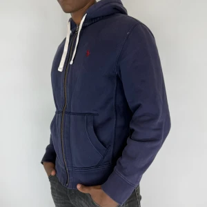 Ralph Lauren Zip Hoodie - Nu har vi nöjet att erbjuda en exklusiv hoodie från det välrenommerade varumärket Ralph Lauren. Hoodien är i storlek Medium och är i mycket gott skick, utan några synliga defekter. Modellen på bilden är 180 cm lång och väger 75 kg, vilket kan ge en god indikation på plaggets passform.
