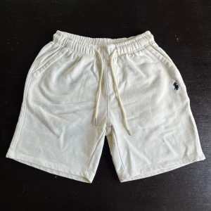 Vita shorts från Polo Ralph Lauren - Snygga vita shorts från Polo Ralph Lauren med klassisk broderad logga i mörkblått på vänster ben. Shortsen har elastisk midja med snörning, sidofickor och en bakficka. Perfekta för sommardagar och avslappnad stil.