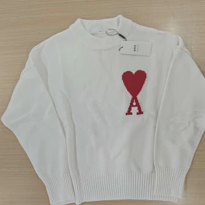 Vit stickad tröja med rött hjärta från Ami - Vit stickad tröja från Ami med ett rött hjärta och bokstaven A broderat på bröstet. Tröjan har rund halsringning, långa ärmar och ribbade muddar vid ärmslut och nederkant. Perfekt för en stilren och trendig look.