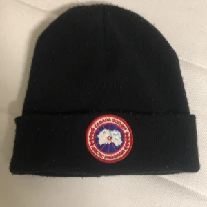 Svart mössa från Canada Goose - Svart mössa med uppvikt kant och klassisk Canada Goose-logga framtill. Enkel och stilren design som passar perfekt till kalla dagar. Tillverkad i mjukt material för extra komfort.