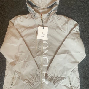 Moncler windbreaker  - Säljer en grå windbreaker från Moncler med luva. Jackan har Moncler-loggan på sidan. Perfekt nu till sommaren eller till hösten! Stort Moncler-tryck längs dragkedjan framtill. Storlek M(kan passa S). Nyskick. Skriv för frågor!🚀