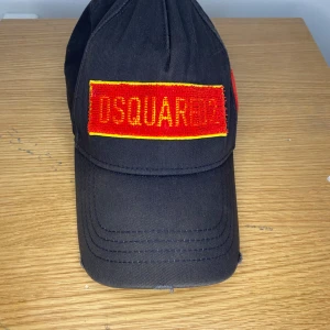 Svart keps från Dsquared2 med röd brodyr - Svart keps från Dsquared2 med röd och gul broderad patch framtill och på sidan. Justerbar baktill med metallspänne. Märkets logga och texten 'Dean & Dan Caten' broderat i rött baktill. Klassisk böjd skärm.