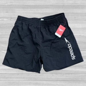 Speedo badshorts - Svarta badshorts från Speedo med vitt logotryck längs ena benet. Klassisk modell med resår i midjan och snörning på insidan. Tillverkade i 100% polyester, perfekta för simning och strandliv.