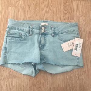 Ljusblå jeansshorts från Nelly - Snygga ljusblå jeansshorts från Nelly med  kanter och dubbla knappar framtill. Shortsen har bakfickor med dekorativt broderi i rosa och vitt. Perfekta för sommaren! 