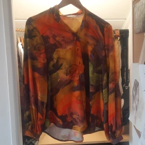 Färgstark blus från Tatuum - Säljer en färgstark och luftig blus från Tatuum med abstrakt mönster i orange, rött, gult, grönt och svart. Blusen har långa ärmar med knappar vid ärmslut och klassisk krage. Perfekt för dig som gillar unika plagg med mycket färg. Vintage/retro.
