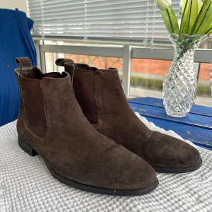 Bruna chelsea boots i mocka - Säljer ett par klassiska bruna chelsea boots i mocka med elastiska paneler på sidorna och låg klack. Perfekta för dig som gillar stilrena och tidlösa skor. Passar till både jeans och kostym.
