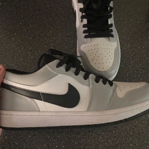 Nike Air Jordan 1 Low i vitt och grått - Säljer ett par Nike Air Jordan 1 Low i vitt och ljusgrått med svarta detaljer och svart swoosh. Skorna har svarta skosnören och perforerad tå för extra ventilation. Klassisk och stilren modell som passar till mycket. Använd fåtal gånger. 