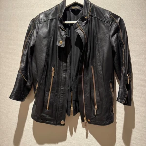 Svart skinnjacka Roberta Biagi - Snygg svart skinnjacka från Roberta Biagi med guldiga dragkedjor och coola detaljer längs ärmar och sidor. Jackan har flera fickor med dragkedja och en stilren, figurnära passform. Perfekt för dig som vill ha en edgy look med exklusiv känsla.