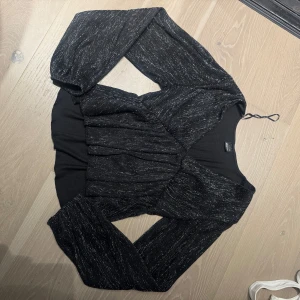 Svart glittrig topp från Gina Tricot - Säljer en svart glittrig topp från Gina Tricot med långa ärmar och lätt puff vid axlarna. Toppen har en figurnära passform och ett snyggt skimmer i tyget. Perfekt för att sticka ut lite extra!