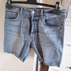 Bermuda shorts i denim med hög midja - Säljer ett par klassiska blå bermuda shorts i jeans med hög midja. Modellen har råa kanter nertill och knappgylf. Perfekta för sommaren och enkla att matcha med olika toppar.