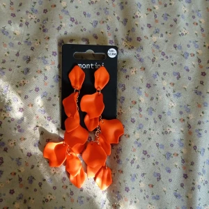 Orange örhängen från Montini/ur&penn - Statementörhängen i orange med stora, böljande blombladsliknande delar. Hängande modell som ger en iögonfallande look. Perfekta för att liva upp en outfit med färg och form.Från ur&penn. Vet inte om det är nyckelfritt.