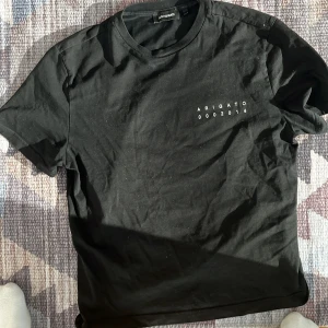 Svart t-shirt från Axel Arigato med fågelmotiv - Svart t-shirt från Axel Arigato med tryck 'ARIGATO 0002014' på bröstet och en stor färgglad fågel på ryggen. Klassisk rund hals och korta ärmar. Perfekt för en stilren och cool look.