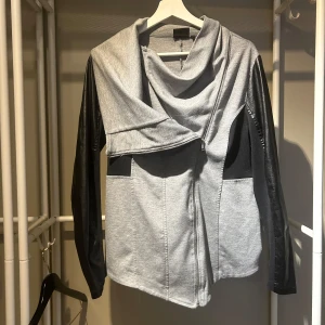 Grå jacka med svarta ärmar och stor krage - Säljer en grå jacka med svarta ärmar och cool asymmetrisk dragkedja. Jackan har en stor, draperad krage och två fickor framtill. Ärmarna är i ett material som liknar skinn, vilket ger en snygg kontrast till den grå kroppen. Den är lite längre i fram än bak. 