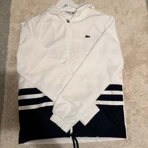 Vit och marinblå vindjacka från Lacoste - Snygg vit vindjacka från Lacoste med marinblå detaljer och ränder nertill.                             Jackan är storlek M, sällsynt vindjacka som är svår att hitta i hemsidan. Pris kan diskuteras vid snabba affärer.        