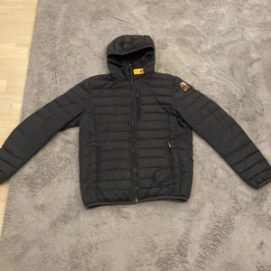 Svart pufferjacka från Parajumpers - Säljer en svart pufferjacka från Parajumpers med huva och dragkedja. Jackan har tre fickor med dragkedja framtill och en patch med logga på ena ärmen. Perfekt för kyliga dagar. Det finns ett litet hål som knappt syns på armen men det kommer ut lite ull då och då.
