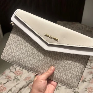 Vit och beige kuvertväska från Michael Kors - Snygg kuvertväska från Michael Kors i en stilren kombination av vitt och beige. Väskan har ett diskret mönster och en elegant logotyp i guld på framsidan. Perfekt för att bära det viktigaste med stil. Aldrig använd. 
