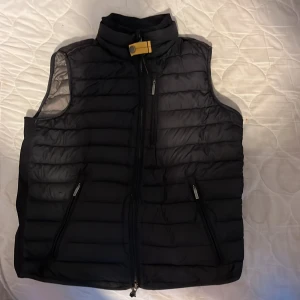 Svart dunväst från Parajumpers - Säljer en svart dunväst från Parajumpers med ståkrage och dragkedja framtill. Västen har två snedställda fickor med dragkedja och en gul detalj vid halsen. Perfekt för lager-på-lager och stilren look.