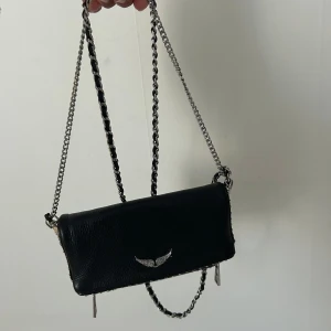 Svart axelväska från Zadig & Voltaire - Säljer en svart axelväska från Zadig & Voltaire med silverfärgad kedjerem och en snygg vingdetalj i metall framtill. Väskan är i skinn och har en stilren, avlång form. Ny pris 2000-3000kr. 