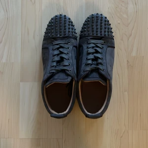 Grå Christian Louboutins - Säljer ett par svarta sneakers från Christian Louboutin med nitar och röd sula. Skorna har snörning och mocha detaljer. Kommer med originalkartong och dustbag. Pris kan diskuteras