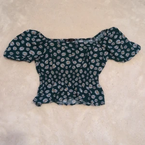 Blommig topp med puffärmar från Sinsay - Supersöt mörkgrön topp från Sinsay med vita och gula blommor. Toppen har puffiga korta ärmar, smockad midja och en croppad passform. Perfekt för dig som gillar romantisk stil och blommiga mönster.