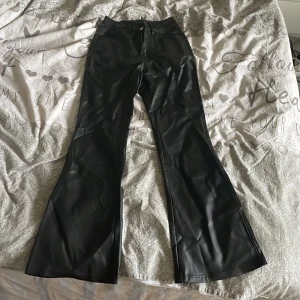 Svarta bootcut byxor i skinnimitation - Säljer ett par svarta bootcut skin-byxor i skinnimitation med mellan midja. Byxorna har aldrig andvänds  och dragkedja framtill och är vida nertill för en snygg siluett. Perfekta för dig som vill sticka ut med en trendig look.
