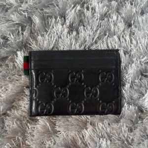 Svart korthållare från Gucci - Säljer en svart korthållare från Gucci i skinn med präglad GG-logga över hela framsidan. Klassisk design med plats för flera kort och en liten detalj i grönt och rött på sidan. Perfekt accessoar för dig som vill ha något stilrent och lyxigt.