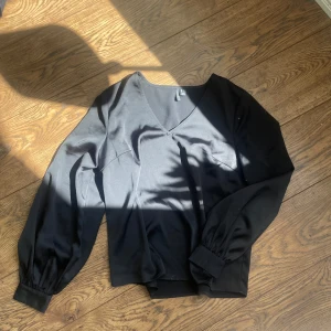 Svart v-ringad blus med ballongärmar - Säljer en svart blus med v-ringning och långa ballongärmar. Blusen har breda muddar vid ärmslut och midja, vilket ger en snygg siluett. Perfekt att styla till både jeans och kjol.