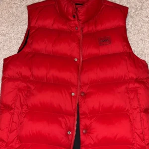 Röd dunväst från Hugo - Snygg röd dunväst från Hugo med hög krage och tryckknappar framtill. Funkar till vår, sommar men även kyligare dagar. Använd väldigt få gånger då den är stor för mig. Ordinarie pris ca 3000, köpt på rea i butik för 2199. 