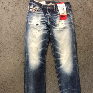 Blå slitna jeans från Diesel - Säljer ett par blå jeans från Diesel med slitningar och ljusa tvättade partier. Jeansen har klassisk femficksdesign, knappgylf och tydlig Diesel-logga bak i midjan. Snyggt distressed look och italiensk design. Den är även i bra skick, inte använd så mycket 