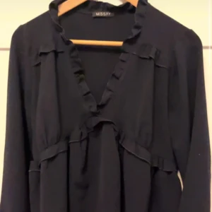 Mörkblå volangblus från Missay - Mörkblå blus från Missay med långa ärmar och volangdetaljer längs urringningen och över bysten. Blusen har en mjuk, flytande passform och är perfekt för dig som gillar en stilren look med lite extra detalj.