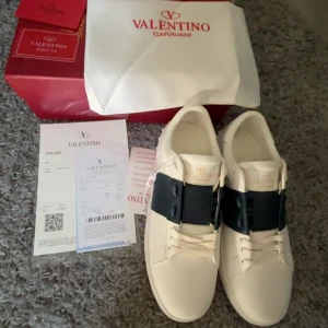Valentino Garavani Open sneakers med blå detalj - Säljer ett par vita Valentino Garavani Open sneakers med bred mörkblå rem över mitten. Skorna har rund tå, platt sula och är tillverkade i skinn. Klassisk och stilren design med subtila nitar längs sulan. Kommer med originalkartong och dustbag.