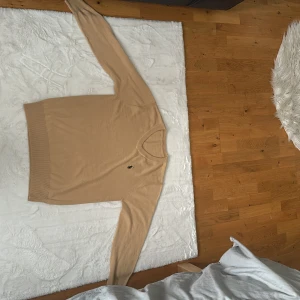 Beige stickad tröja från Ralph Lauren - Snygg beige stickad tröja från Ralph Lauren med klassisk v-ringning och broderad logga på bröstet. Perfekt för en stilren och avslappnad look. Långa ärmar och ribbade muddar vid ärmslut och nederkant.