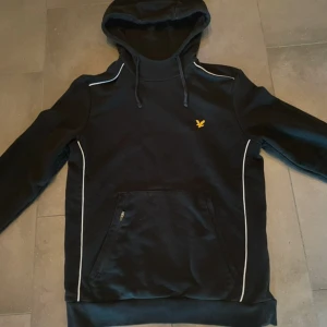 Svart hoodie från Lyle & Scott med vita detaljer - Säljer en svart hoodie från Lyle & Scott med vita detaljer och en gul logotyp på bröstet. Tröjan har en praktisk ficka med dragkedja framtill och en justerbar huva. Perfekt för en stilren och bekväm look. Helt oanvänd! Nypris:899kr, bara höra av sig vid frågor!