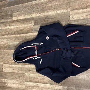 Mörkblå Moncler hoodie med röd/vita detaljer - Snygg mörkblå hoodie från Moncler med dragkedja och huva. Tröjan har vita och röda detaljer längs dragkedjan, fickorna och huvan. Klassisk Moncler-logga på bröstet och vita snören vid huvan. Perfekt för dig som gillar streetwear med premiumkänsla.