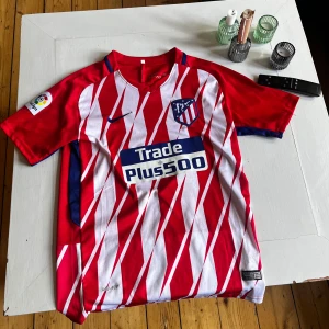 Atletico Madrid matchtröja Nike röd/vit - Säljer en Atletico Madrid fotbollströja från Nike med röda och vita diagonala ränder, klubbmärke på bröstet och La Liga-märke på ärmen. Tröjan har blå detaljer, korta ärmar och är tillverkad i lätt funktionsmaterial. Perfekt för dig som älskar spansk fotboll.