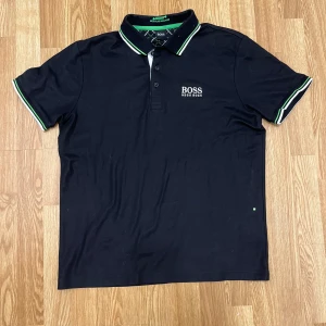 Svart pikétröja från Hugo Boss - Snygg svart pikétröja från Hugo Boss med vit och grön rand på krage och ärmslut. Klassisk polokrage med knappar och broderad logga på bröstet. Tillverkad i bomull för en skön och luftig känsla. Perfekt för en stilren och sportig look.