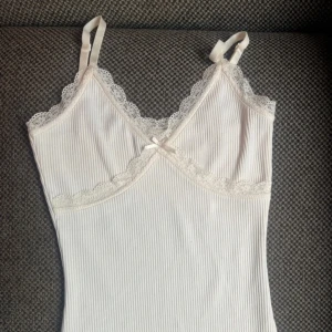 Vit ribbad spetsklänning med rosett - Supersöt kort vit klänning med tunna axelband, ribbat tyg och spetsdetaljer längs urringningen och under bysten. Liten rosett framtill för extra gullig touch. Perfekt för sommarens varma dagar eller när du vill känna dig extra fin.