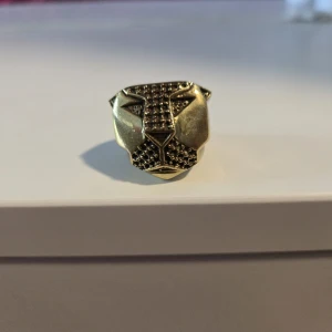 Guldig ring med pantermotiv - Cool ring i guldtonad metall med ett detaljerat pantermotiv framtill. Ringen har en robust och chunky design med markerade drag och texturerad yta på panterns ansikte. Perfekt statement piece för dig som gillar djur och unika accessoarer.