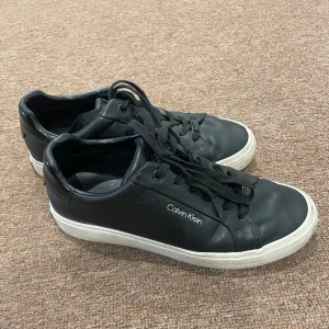 Svarta sneakers från Calvin Klein - Stilrena svarta sneakers från Calvin Klein med vit sula och diskret logga på sidan och hälen. Skorna har snörning och är tillverkade i skinn med klassisk rund tå. Perfekta för dig som gillar en clean och modern look. Skorna har använts ett fåtal gånger, väldigt bra skick.