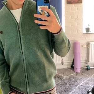 Grön stickad kofta med dragkedja - Snygg grön stickad kofta med dragkedja och ribbade muddar. Har en liten broderad logga på bröstet och diskreta ränder i vitt och rött vid krage och nederkant. Perfekt för lager-på-lager och chill vardagsstil.