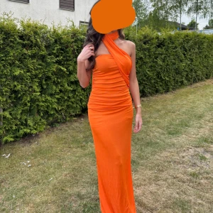 Orange lång klänning med omlott-hals - Säljer en snygg orange långklänning med draperad effekt och omlott-hals som går över ena axeln som en scarf och fortsätter ner bakom ryggen. Klänningen är figurnära och har en ärmlös design med en cool asymmetrisk axel. Perfekt för dig som vill sticka ut med färg och stil.