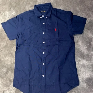 Kortärmad Ralph Lauren Skjorta - Säljer en mörkblå kortärmad skjorta från Ralph Lauren med klassisk krage och vita knappar. Skjortan har en röd broderad logga på bröstet och är tillverkad i bomull. Perfekt för en stilren look. Den är knappt använd och det är bara skriva om ni har några frågor.
