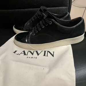 Svarta sneakers från Lanvin med lackad tå - Säljer ett par svarta sneakers från Lanvin med snygg lackad tå och vit sula. Skorna har snörning och är tillverkade i mocka och skinn, vilket ger en lyxig känsla. Perfekta för dig som gillar stilrena och exklusiva sneakers ni inför sommaren!