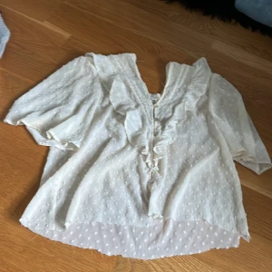 Vit blus med volang från Zara str S - Den är klippt där nere för att den ska bli kortare, därav priset!! Supersöt vit blus från Zara med volangdetaljer längs knäppningen och små prickar i tyget. Blusen har korta ärmar, knappar framtill och en luftig, lätt transparent känsla. Perfekt för dig som gillar romantisk och feminin stil.