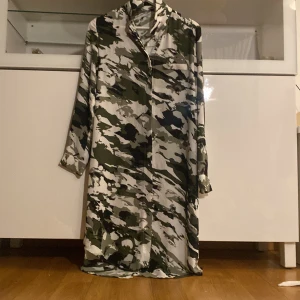 Camouflagefärgad lång skjortklänning - Säljer en lång skjortklänning med camouflage-mönster i grönt, svart, grått och vitt. Klänningen har lång ärm, krage och knäppning framtill. Perfekt för dig som vill sticka ut med en cool och avslappnad stil.