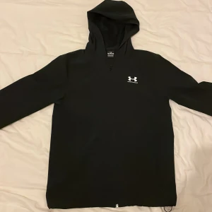 Svart hoodie från Under Armour - Svart hoodie från Under Armour med vit logga på bröstet. Tröjan har huva, lång ärm och en clean design utan extra detaljer. Perfekt för dig som gillar sportig och enkel stil.