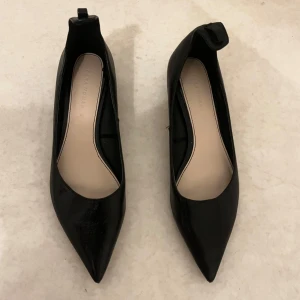 Svarta pumps från Zara Woman - Stilrena svarta pumps från Zara Woman med spetsig tå och låg klack. Skorna är i glansigt material och har en unik detalj baktill på hälen. Perfekta för dig som gillar klassisk och trendig stil.