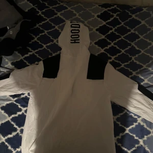 Vit hoodie från Hoodrich med svarta detaljer - Säljer en vit hoodie från Hoodrich med svarta detaljer på axlarna och texten 'HOODRICH.' på bröstet och 'HOOD' på huvan. Stor magficka framtill och justerbar huva. Perfekt för en streetwear-look.