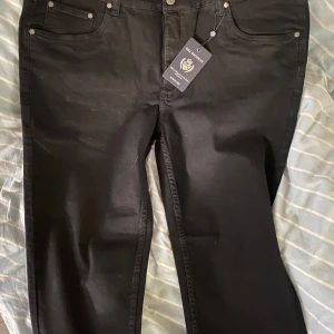 Svarta jeansbyxor från Mel Richman - Säljer ett par svarta jeansbyxor från Mel Richman med klassisk femficksdesign och bälteshällor. Byxorna har normal passform och stängs med knapp och dragkedja. Perfekta för en stilren look.